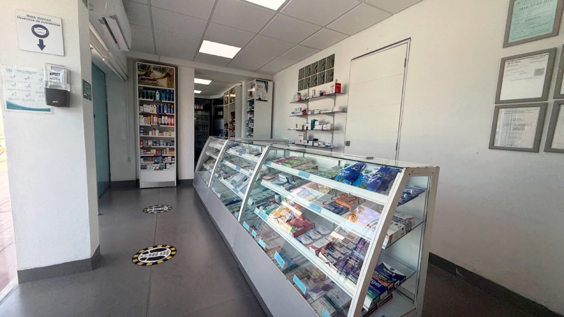El mostrador de la farmacia externa: la vitrina con los productos a la vista, las estanterías del fondo y las licencias enmarcadas en la pared.