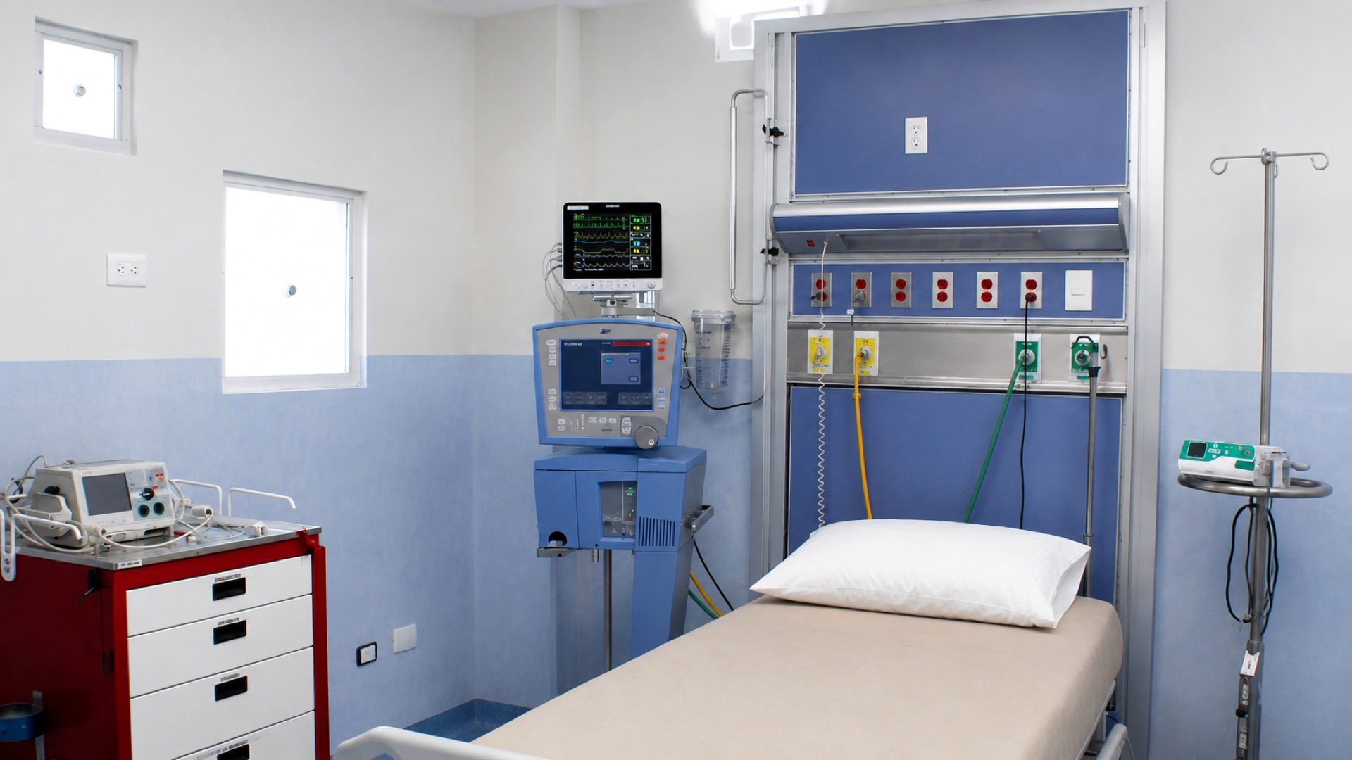 Un puesto de terapia intensiva: la cama contra el panel de cabecera con sus tomas de gases, el ventilador y el monitor de signos vitales a un lado, y el carro de paro al otro.
