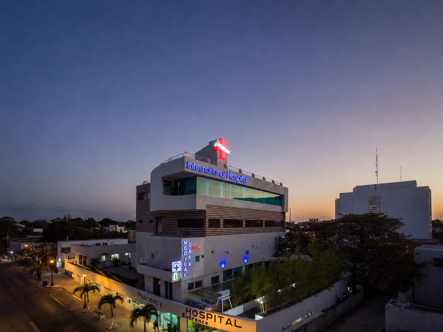 El edificio del hospital al anochecer, visto desde el aire: la cruz roja encendida en la azotea, el rótulo «International Hospital» en neón azul y, a pie de calle, las entradas de la farmacia y del hospital.